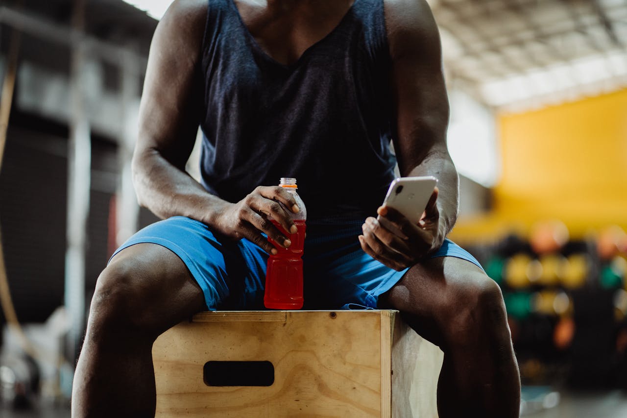 Athlete seated on box holding a smartphone and energy drink, blurred gym background.